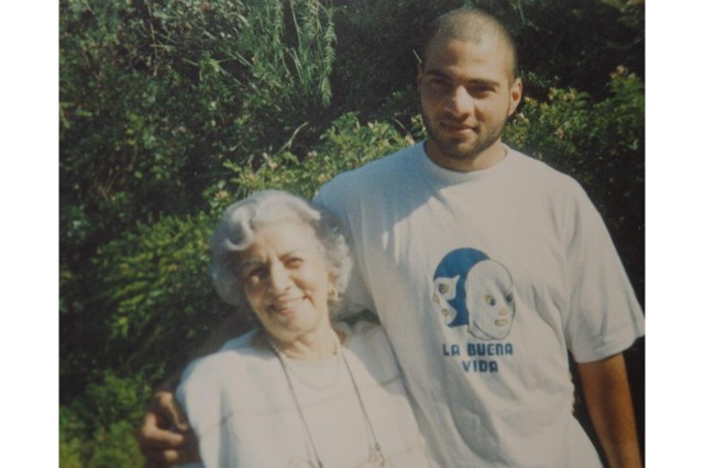 Young man with shaved head, in a white t-shirt with his right arm around his white-haired grandmother who wears a white top and comes up to the man's shoulder.